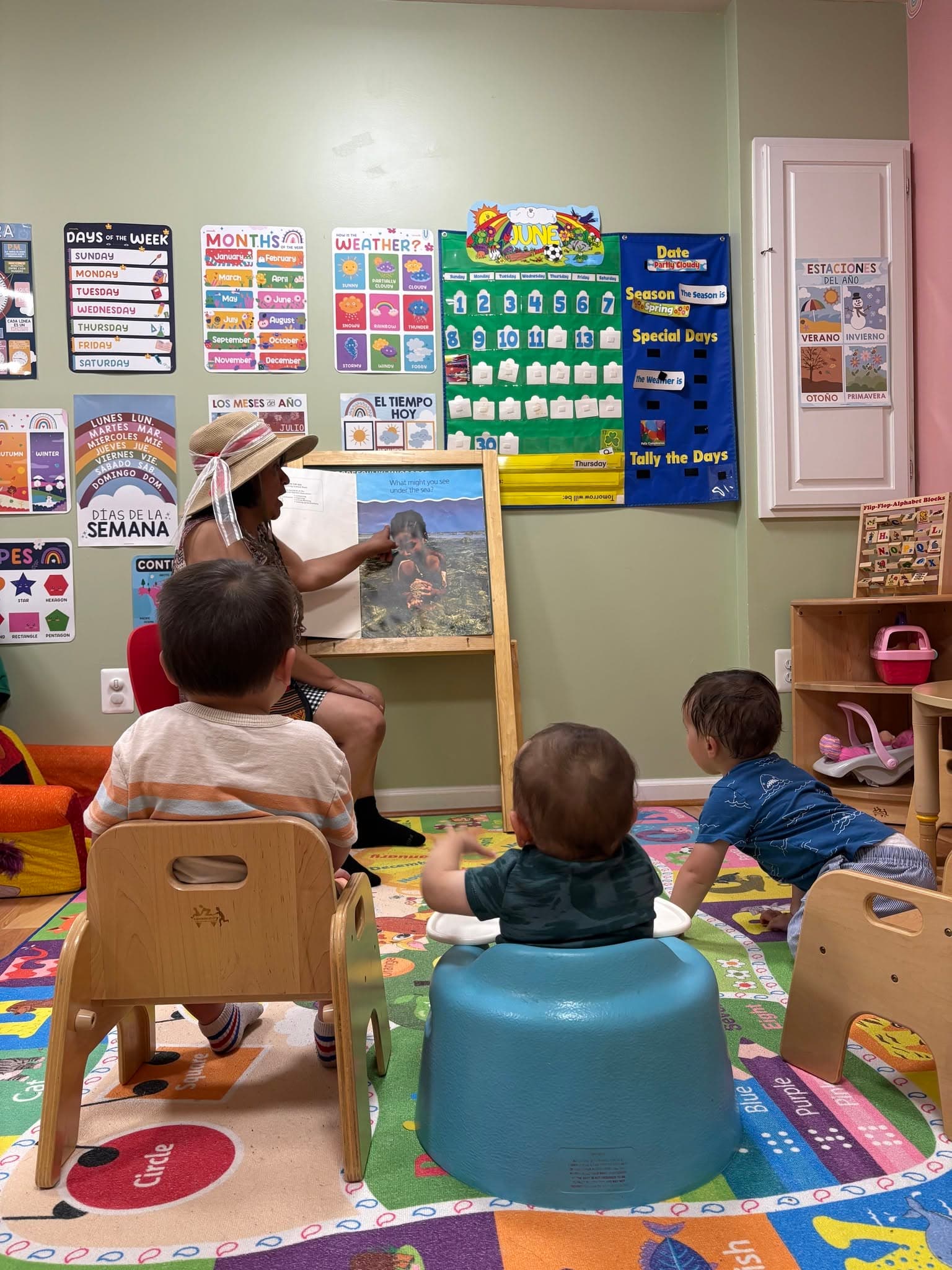 Justina at the easel during circle time, pointing at a picture while three children sit and watch, with days-of-the-week and weather charts in English and Spanish on the wall behind.