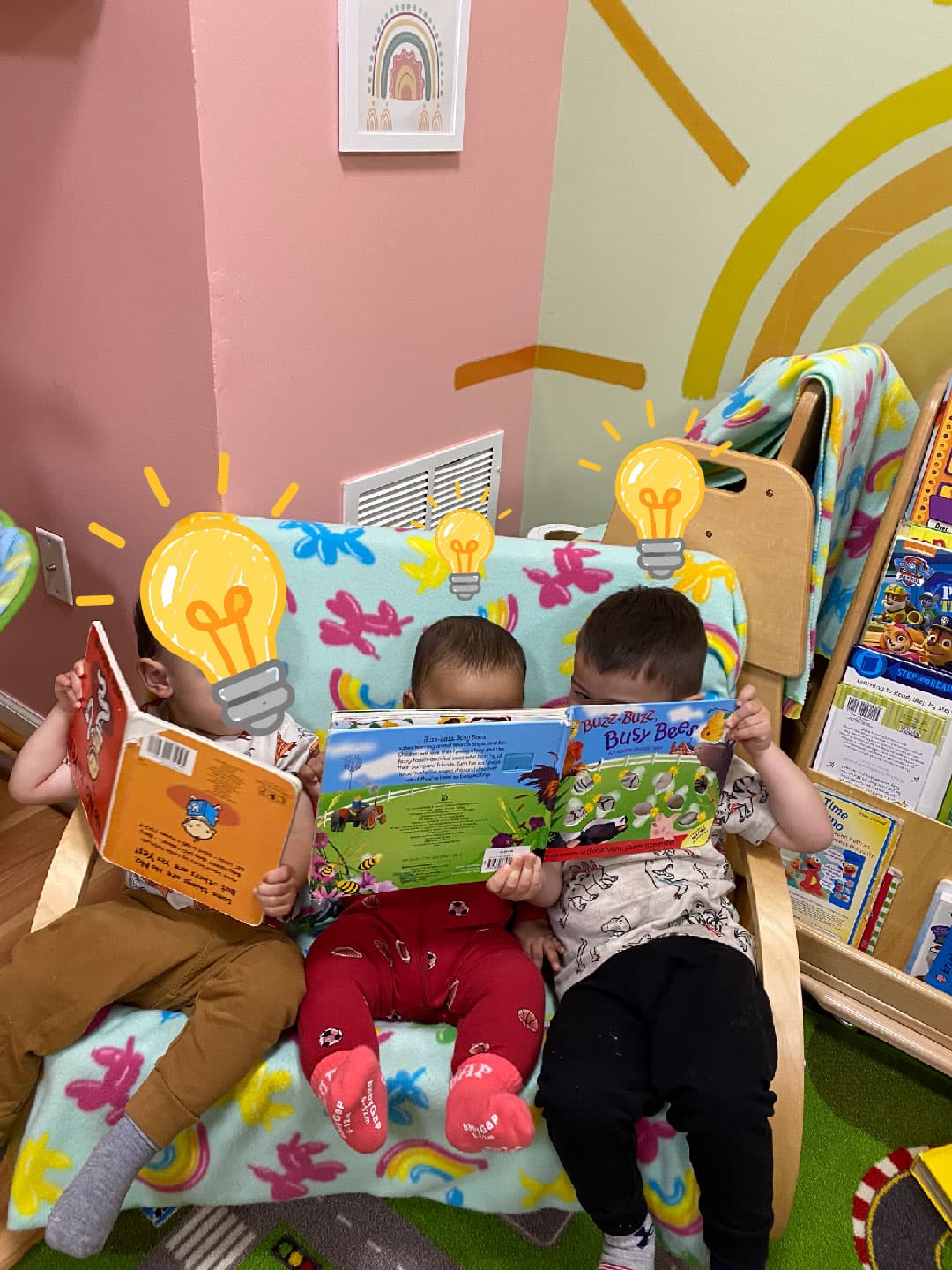 Three toddlers sitting side by side on the couch in the reading corner. The one on the left is holding his book upside down; the other two are reading one book together.