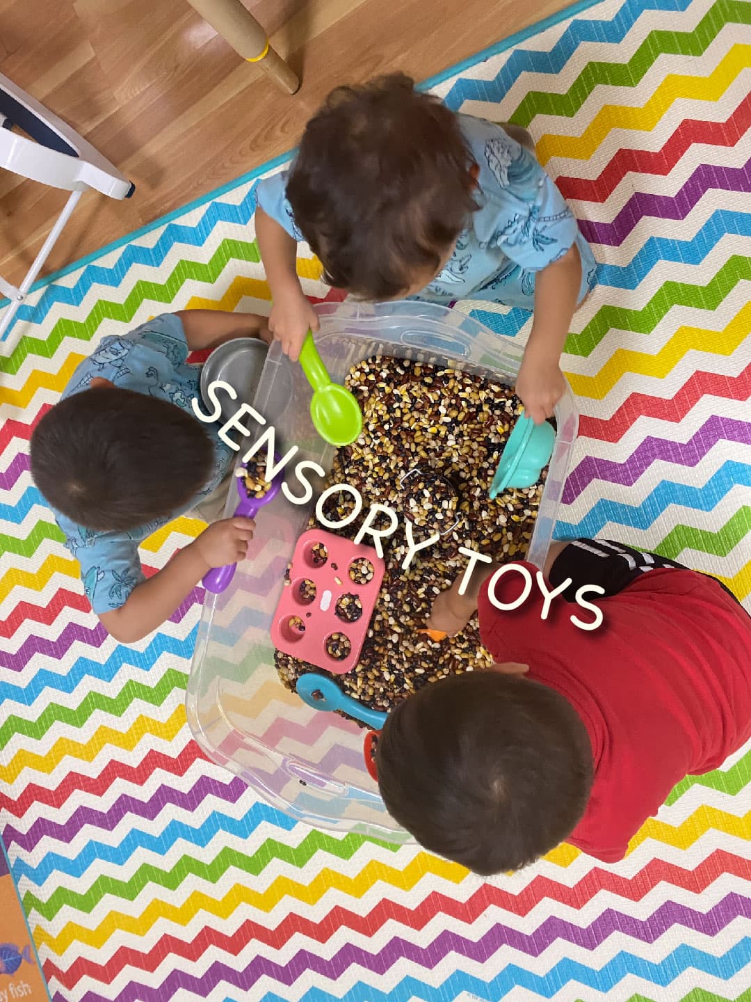 Three children seen from above, scooping dry beans with cups and a muffin tray in a sensory bin on a rainbow rug.