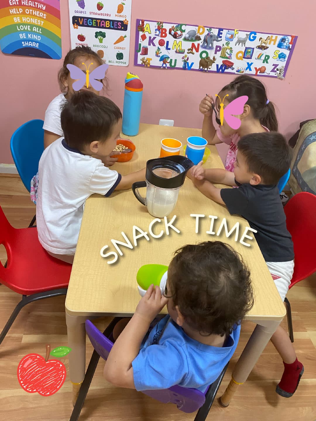 Five children sitting together around a low table at snack time, with cups and bowls in front of them and alphabet charts on the wall behind.