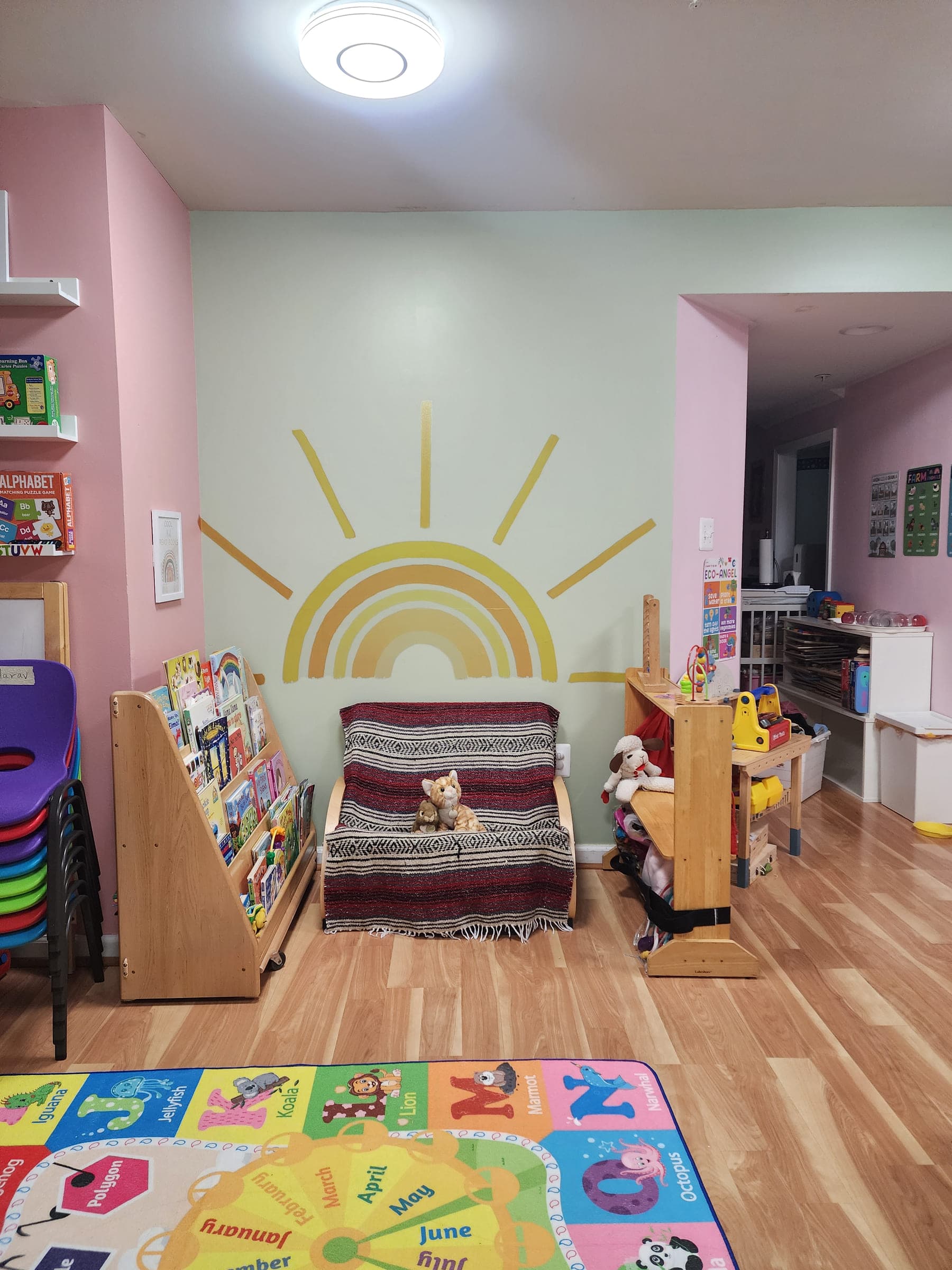 The reading corner, with a sunrise painted on the wall, a small couch, a rack of picture books, and a hallway behind leading to the bathroom and changing area.
